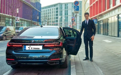 SL-Limousines I Chauffeur & Limousine Service - BMW Luxury Limousine An elegantly dressed chauffeur stands next to a black BMW luxury sedan with an open door.