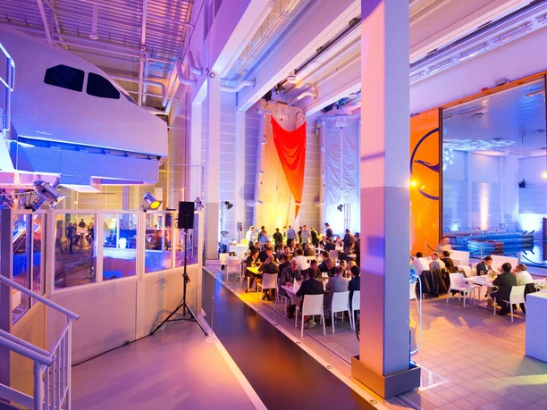 Lufthansa Aviation Training Center Frankfurt - Veranstaltungshalle Ein großer, modern ausgeleuchteter Veranstaltungssaal mit hohen Decken, bunter Beleuchtung und vielen Gästen, die an weißen Tischen sitzen und speisen.