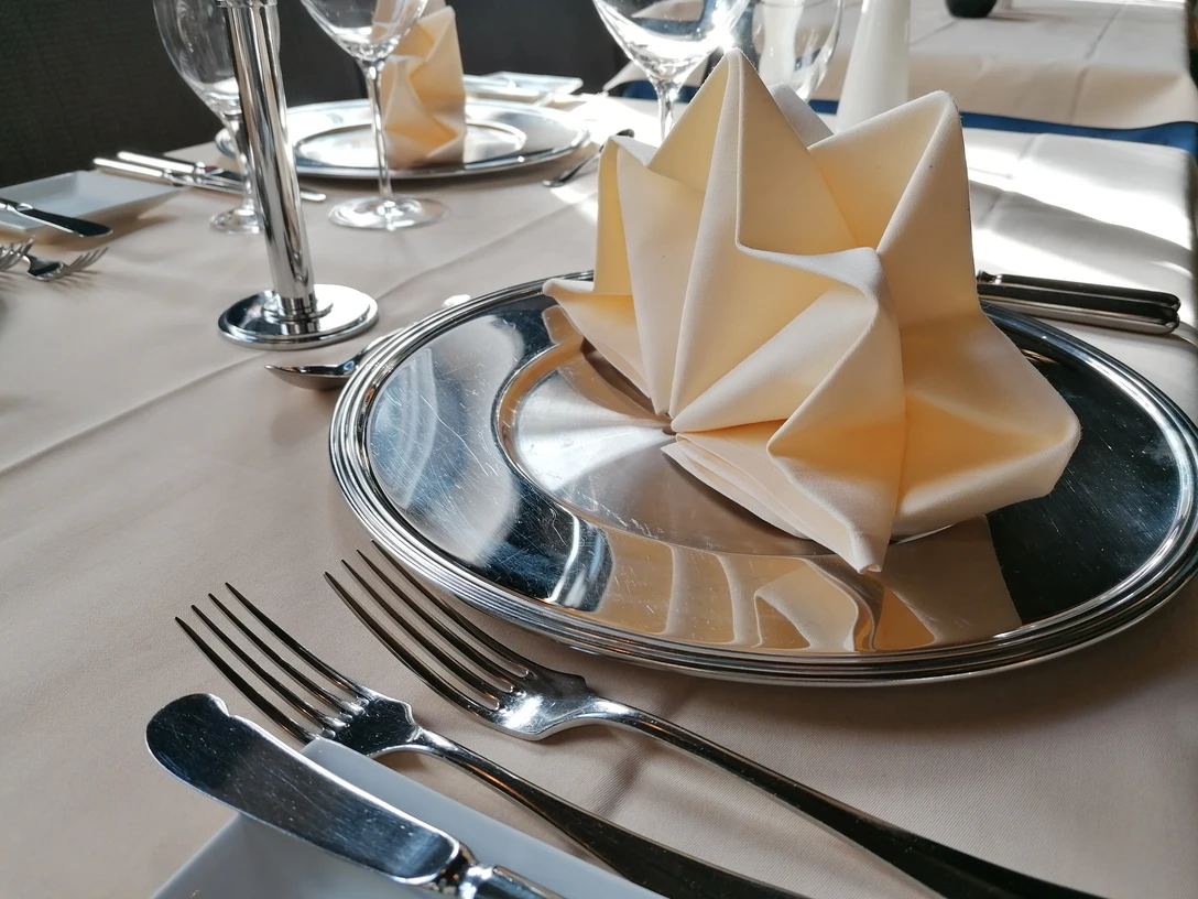 Gedeckter Tisch mit gefalteter Stoffserviette auf Metallteller und silberfarbenem Besteck.