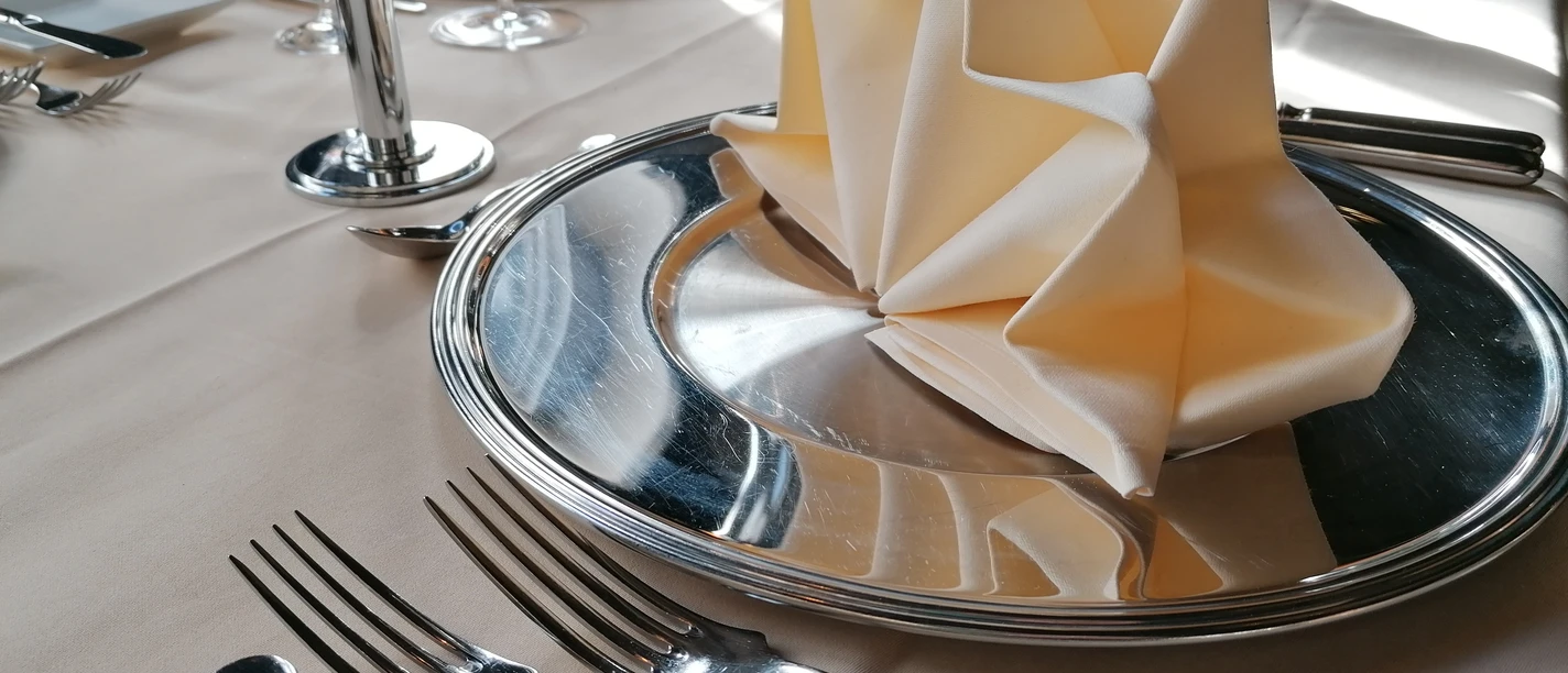 Gastronomie in Beverungen (Beispielfoto) Gedeckter Tisch mit gefalteter Stoffserviette auf Metallteller und silberfarbenem Besteck.