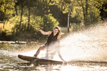 Wakeboarden bei Wasserski Langenfeld Lachende Person auf einem Wakeboard, umgeben von Sonnenlicht, das im Wasser spritzt.