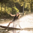 Wakeboarden bei Wasserski Langenfeld Lachende Person auf einem Wakeboard, umgeben von Sonnenlicht, das im Wasser spritzt.