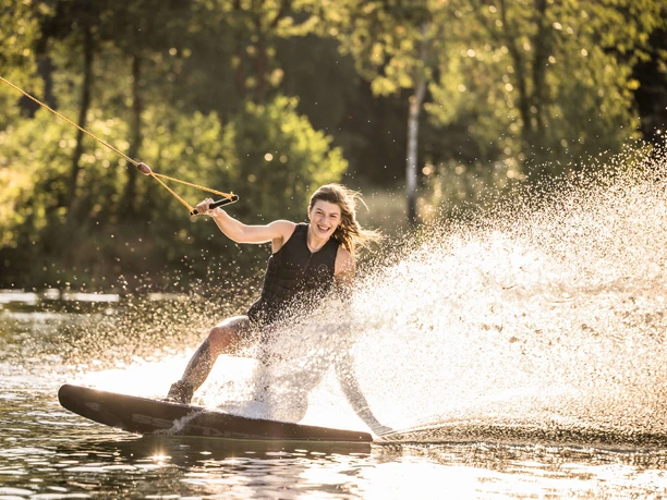 Wakeboarden bei Wasserski Langenfeld Lachende Person auf einem Wakeboard, umgeben von Sonnenlicht, das im Wasser spritzt.