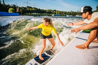 Surferin auf der stehenden Welle bei Wasserski Langenfeld Junge Frau surft auf einer stehenden Welle bei der Wasserskianlage in Langenfeld, ein Mann assistiert vom Rand.