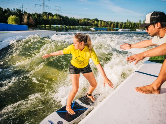 Surferin auf der stehenden Welle bei Wasserski Langenfeld Junge Frau surft auf einer stehenden Welle bei der Wasserskianlage in Langenfeld, ein Mann assistiert vom Rand.