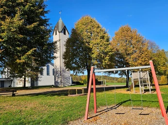 Weißer Kirchturm mit Bäumen und Spielplatz mit Schaukel bei sonnigem Wetter.