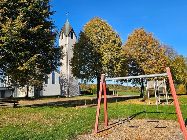 Langeland Nepomuk-Kirche Weißer Kirchturm mit Bäumen und Spielplatz mit Schaukel bei sonnigem Wetter.
