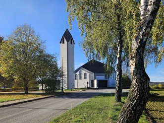 Weißer Kirchturm und Kirchenbau zwischen Bäumen an einer ruhigen Straße bei klarem Himmel.