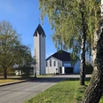 Weißer Kirchturm und Kirchenbau zwischen Bäumen an einer ruhigen Straße bei klarem Himmel.