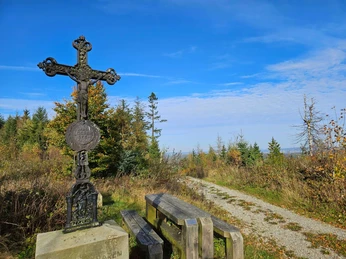 Eisernes Kreuz neben Holzbänken an einem Schotterweg vor herbstlichem Wald und blauem Himmel