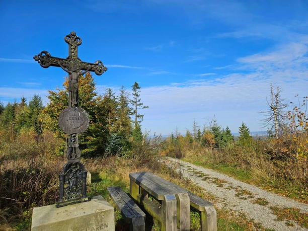Eisernes Kreuz neben Holzbänken an einem Schotterweg vor herbstlichem Wald und blauem Himmel