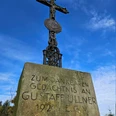 Steinkreuz mit Metallkruzifix und gut lesbarer Widmung vor klarem blauen Himmel.