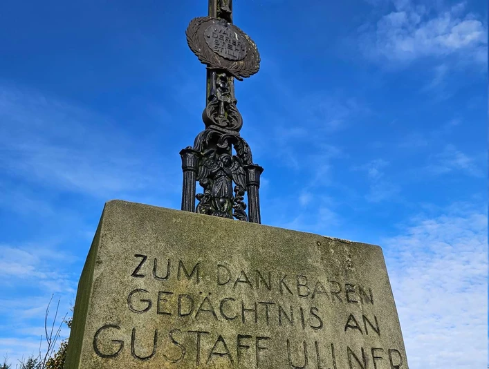 Langeland Schwarzes Kreuz Steinkreuz mit Metallkruzifix und gut lesbarer Widmung vor klarem blauen Himmel.