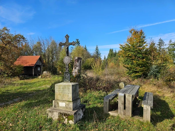 Langeland Schwarzes Kreuz - Hütte Steinkreuz neben Holztisch in einer lichten Waldfläche mit kleiner Hütte im Hintergrund.