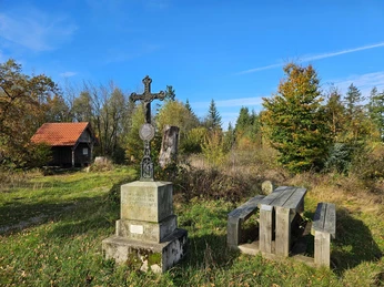 Langeland Schwarzes Kreuz Steinkreuz neben Holztisch in einer lichten Waldfläche mit kleiner Hütte im Hintergrund.
