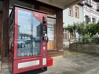 Getränkeautomat vor einer Hauswand, daneben Pflanzen und ein Gebäude.
