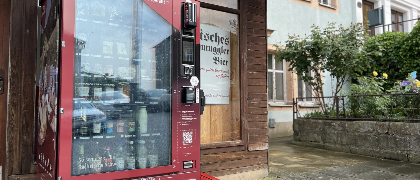 Getränkeautomat vor einer Hauswand, daneben Pflanzen und ein Gebäude.