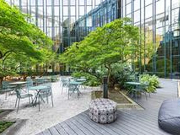 Design Offices Frankfurt Westendcarree - Außenbereich Ruhiger Außenbereich mit hellgrünen Bäumen, hellen Kiesbetten und Sitzgelegenheiten im Schatten eines modernen Glasgebäudes.