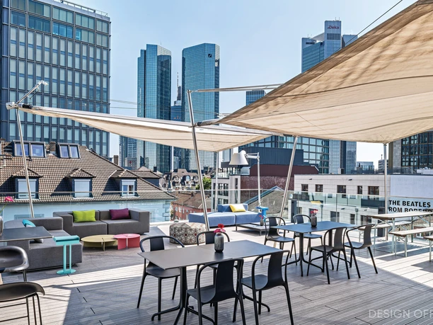 Design Offices Frankfurt Barckhausstraße - Dachterrasse Sonnige Dachterrasse mit Loungemöbeln und Tischen, beschattet von einem Segel. Im Hintergrund dominiert die Skyline von Frankfurt mit modernen Wolkenkratzern.