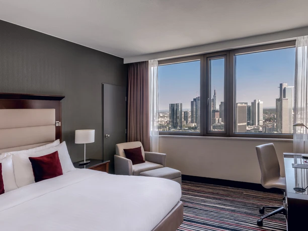 Frankfurt Marriott Hotel - Skyline Room Ein modernes Hotelzimmer mit großem Bett und Schreibtisch. Der Blick aus dem Fenster zeigt die Skyline von Frankfurt bei Tag.