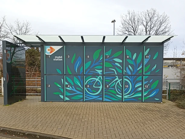 Fahrradboxen mit blau-grünem Blättermotiv und überdachtem Bereich an einem Bahnhof.