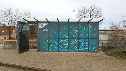 RadboxBahnhofSHS.jpg Fahrradboxen mit blau-grünem Blättermotiv und überdachtem Bereich an einem Bahnhof.