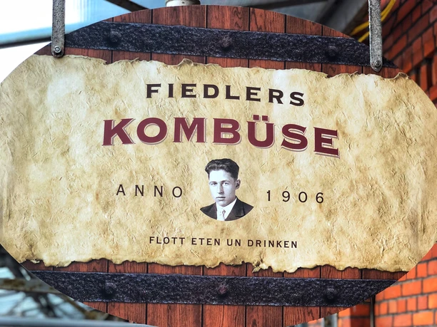 IMG_2618.jpeg Holzschild von Fiedlers Kombüse mit Porträtfoto und Schriftzug über einer Backsteinfassade in Bremerhaven