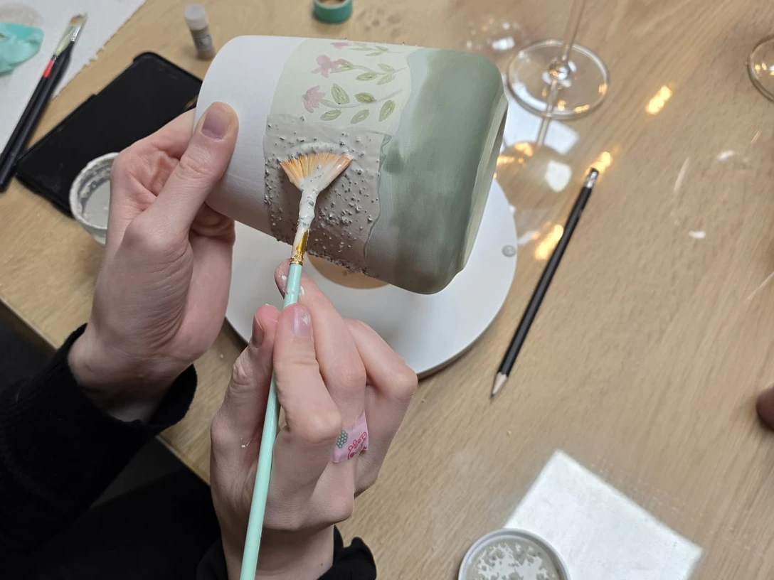 IMG-20251210-WA0008.jpg Person bemalt einen hellgrünen Keramikbecher mit feinem Pinsel und floralen Mustern auf Holztisch