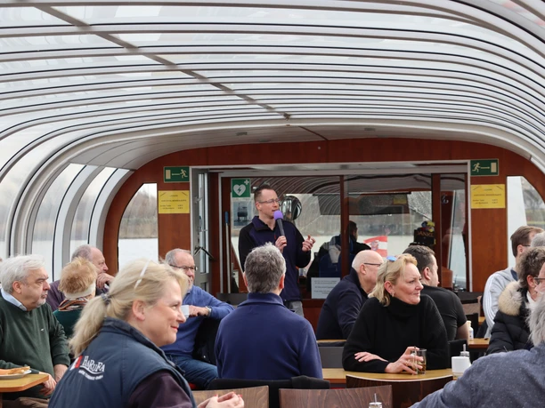 Sprecher 2 (c) Maja Herwig_Erlebnis Bremerhaven.JPG Ein Mann hält ein Mikrofon und spricht vor einer Reisegruppe auf einem verglasten Ausflugsschiff.