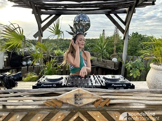 Soundshine Festival, DJane.jpg Eine DJane steht am Mischpult auf einer Bühne aus Holz, im Hintergrund stehen viele Parlmen