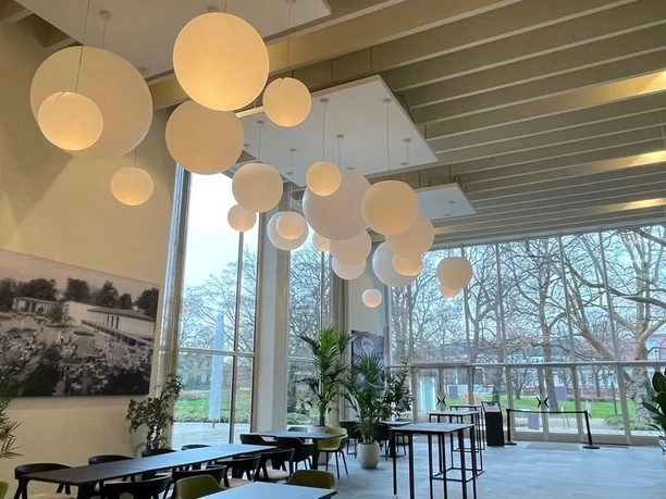 Heller Café-Innenraum mit großen Fenstern, Kugelleuchten, Pflanzen und Blick in den Kurpark
