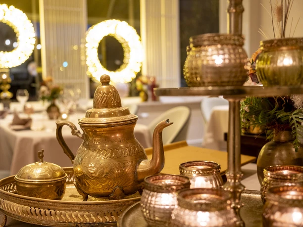 Sahila Eine orientalische goldene Teekanne. Im Hintergrund ein elegant gedeckter Tisch.