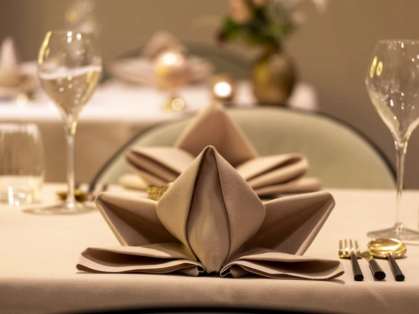 Sahila An elaborately folded beige cloth napkin. Gold cutlery next to it.
