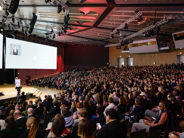 Congress Center Messe Frankfurt - Congress Hall A large crowd sits in a spacious congress hall and looks at a stage on which a speaker is standing and whose picture is shown on a large screen.