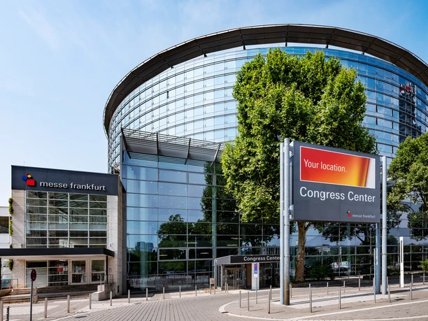 Congress Messe Frankfurt The Congress Center of Messe Frankfurt by day. The modern, round glass façade and the entrance building are visible under a blue sky.