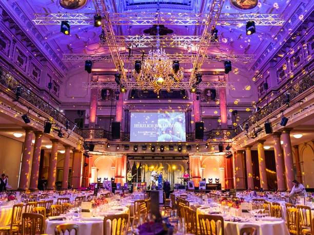 Gesellschaftshaus Palmengarten - Banquet hall An opulently decorated, festive banquet hall with long rows of tables, illuminated in shades of purple and gold, with an orchestra on a stage in the background.
