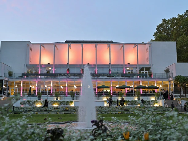 Gesellschaftshaus Palmengarten - illuminated Gesellschaftshaus Das hell erleuchtete Gesellschaftshaus Palmengarten bei Dämmerung, mit einem Springbrunnen und Pflanzen im verschwommenen Vordergrund.