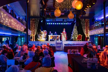 Afternoon trip during Advent Festive hall with a costumed figure on stage, bright lights, decorations and seated guests.