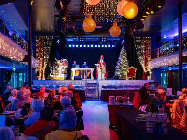 Afternoon trip during Advent Festive hall with a costumed figure on stage, bright lights, decorations and seated guests.