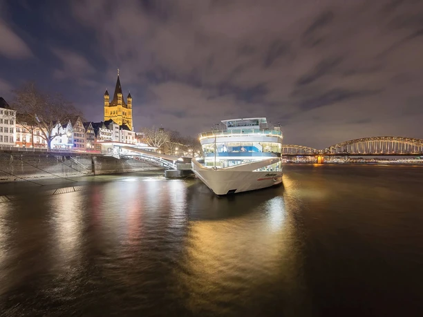 Christmas all-inclusive evening cruise Night view of a lit river cruise ship by waterfront homes and a church beside an arched bridge.