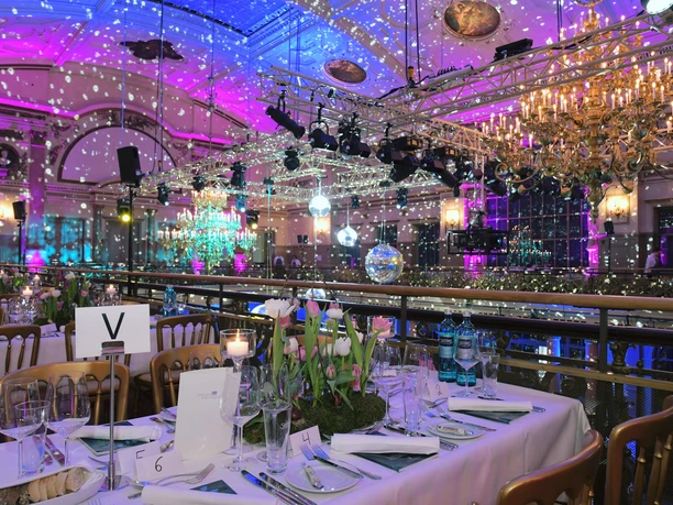 Gesellschaftshaus Palmengarten - Bankettsaal Ein opulent dekorierter Bankettsaal bei Nacht, mit einem festlich gedeckten Tisch im Vordergrund und einer Balustrade, beleuchtet in hellem Magenta und Blau.
