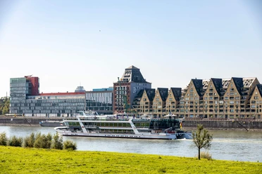 Rheinauhafen Moderne Uferbauten am Rhein mit vorbeifahrendem Ausflugsschiff und grüner Wiese im Vordergrund.
