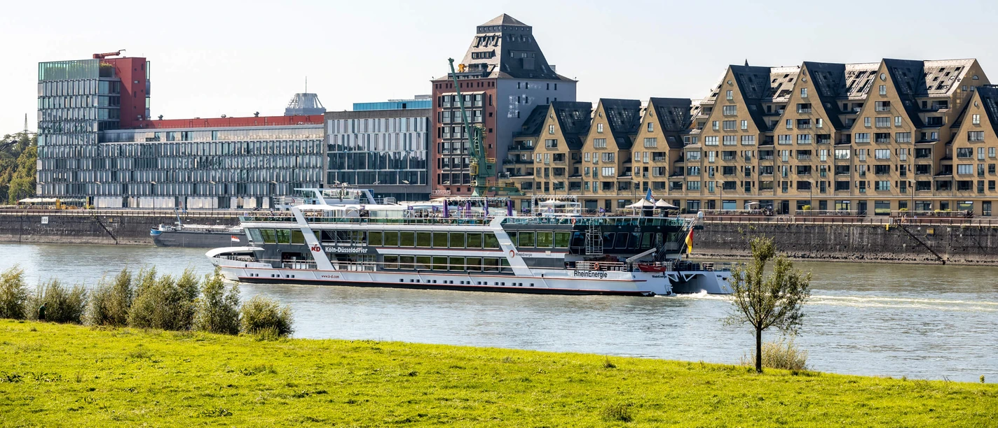 Rheinauhafen Moderne Uferbauten am Rhein mit vorbeifahrendem Ausflugsschiff und grüner Wiese im Vordergrund.