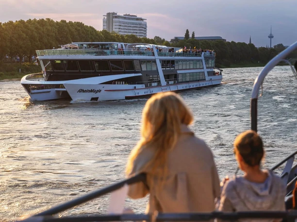 Lounge Abendfahrt Ein beleuchtetes Rheinschiff fährt bei Abendlicht vorbei, während Passagiere vom Deck zusehen.