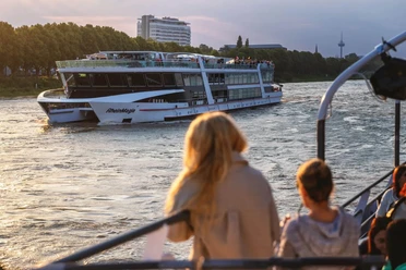 Lounge evening trip An illuminated Rhine ship sails past in the evening light while passengers watch from the deck.