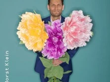 Person in blauem Anzug hält drei übergroße bunte Blumen vor türkisfarbenem Hintergrund.