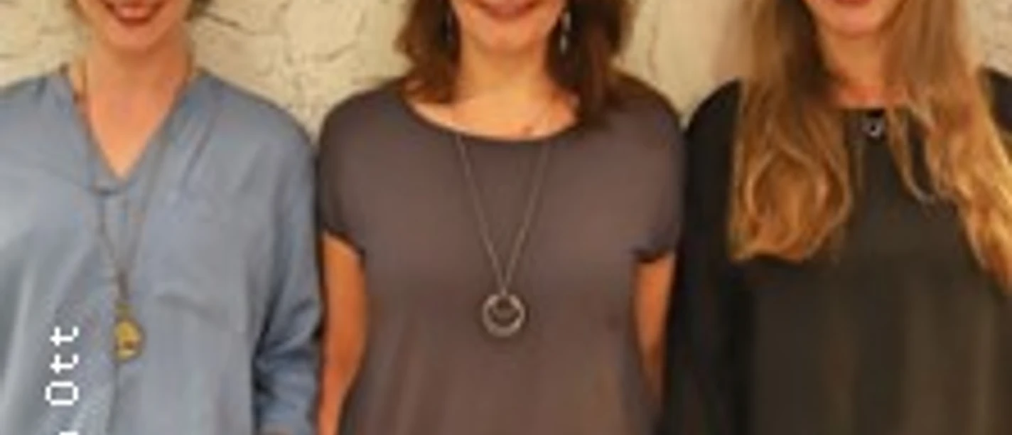 Drei Frauen stehen nebeneinander vor einer hellen Wand und lächeln in die Kamera.