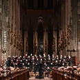 Chor und Orchester vor hoher gotischer Kulisse im Dom, umgeben von beleuchteten Säulen.