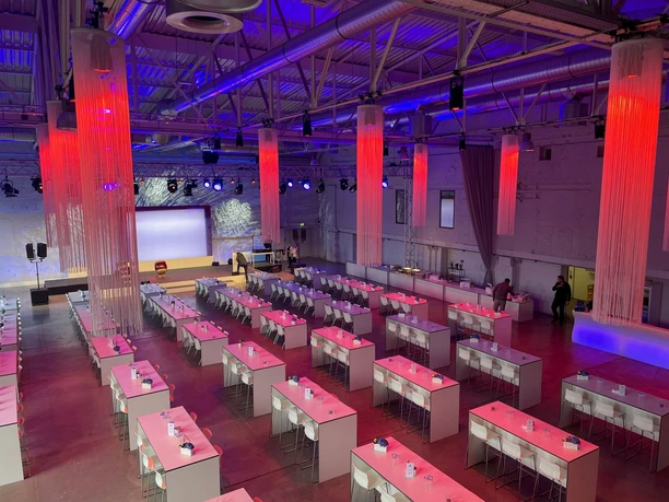 Dock² Eventlocation große Veranstaltungshalle in rotem Licht mit Tischen, Stühlen und Bühne mit Leinwand