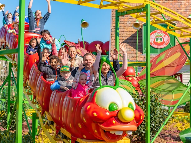 Zdjęcie przedstawia tor gąsienicowy w parku rozrywki Karls Erlebnisdorf. Kilkoro dzieci siedzi w kolorowych wagonikach małej kolejki górskiej i wiwatuje z radości.